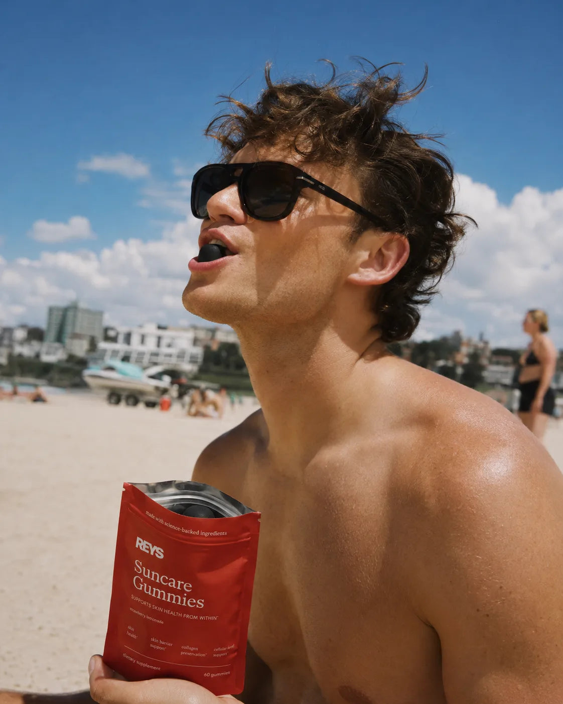 A man at the beach in sunglasses chewing a REYS Suncare Gummy, holding the poppy-red pouch in the midday sun