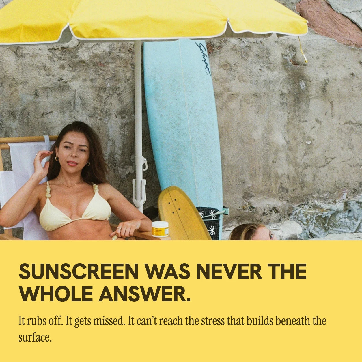 Sunscreen was never the whole answer — it rubs off, gets missed, and can’t reach the stress that builds beneath the surface; two friends under a yellow beach umbrella with the REYS jar